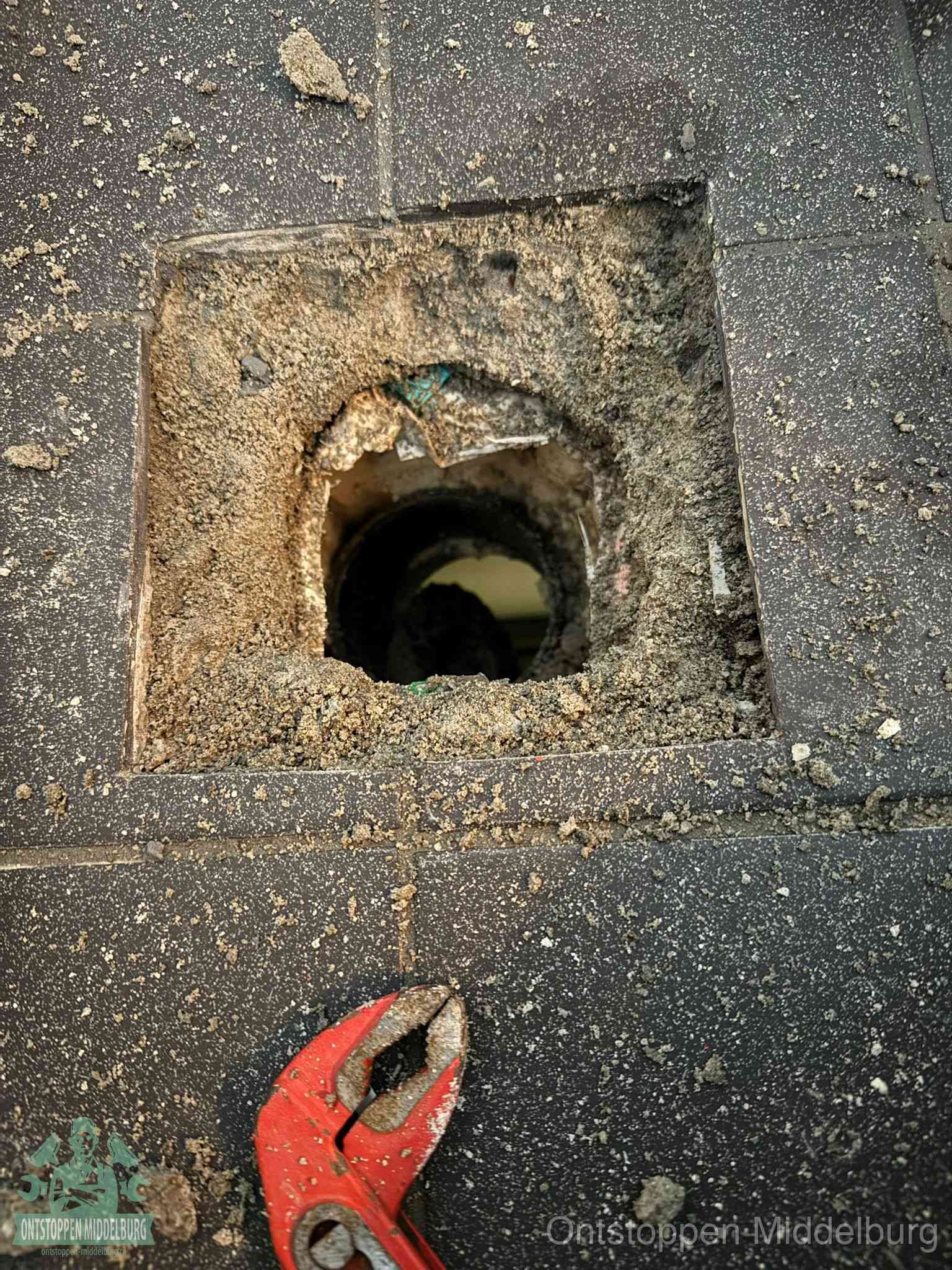 Een geopend rioolputje in een betontegelvloer met losgemaakt rooster en een zichtbare ronde afvoerbuis, het achterhalen waar de afvoerverstopping zit in Middelburg