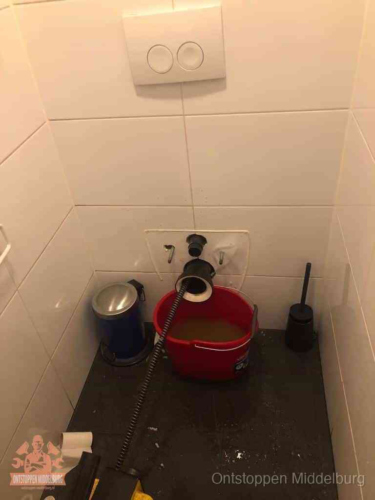 Een ontstopte wand-wc met verwijderde toiletpot waar een ontstoppingsveer in de wanduitgang verdwijnt, het achterhalen waar de verstopping zit in Middelburg