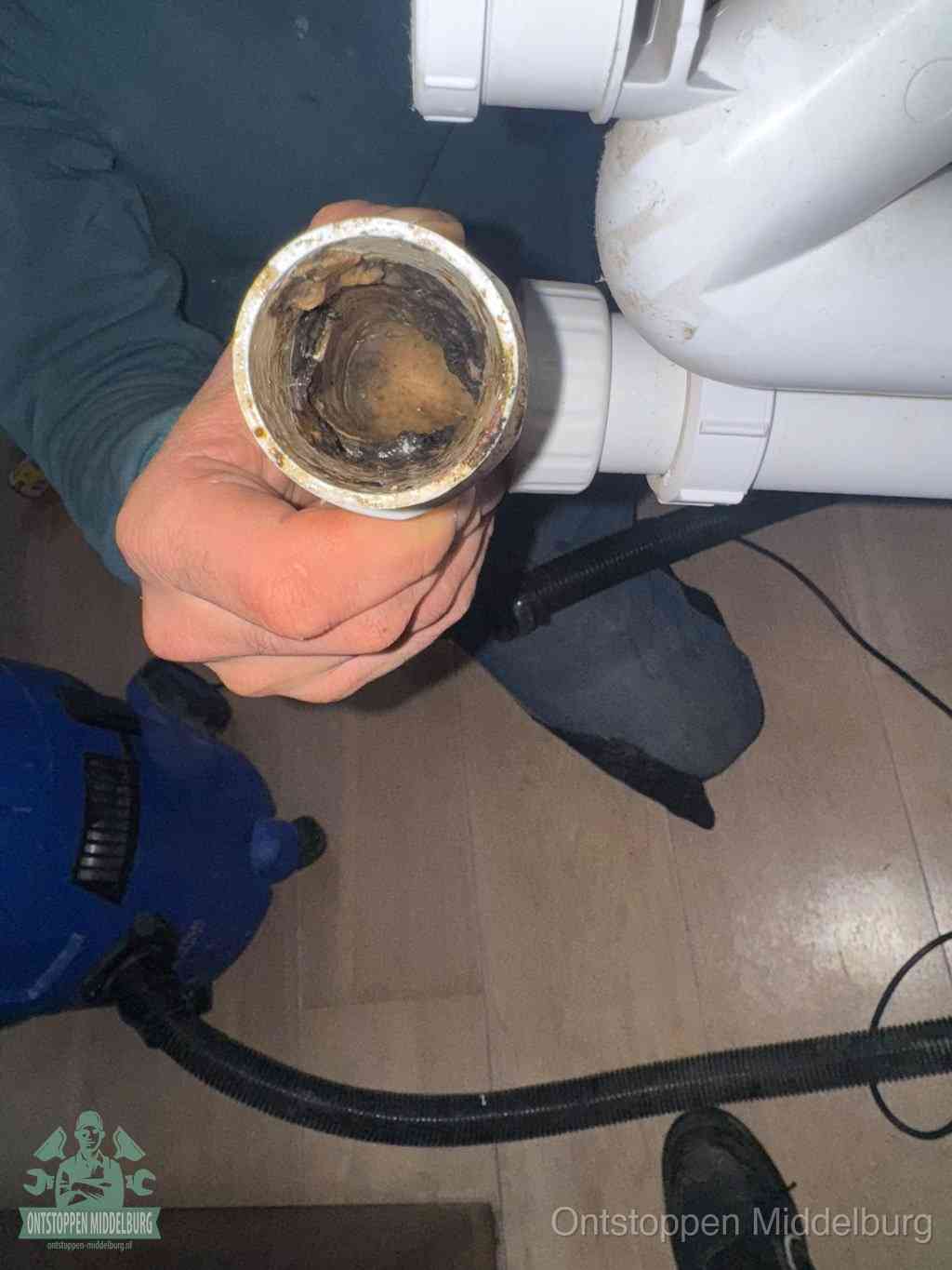Een hand houdt een losgekoppeld PVC-afvoerbuisstuk vast dat van binnen volledig is dichtgeslibd met vet, vuil en aanslag, de meest voorkomende oorzaak van een verstopte afvoer in Middelburg