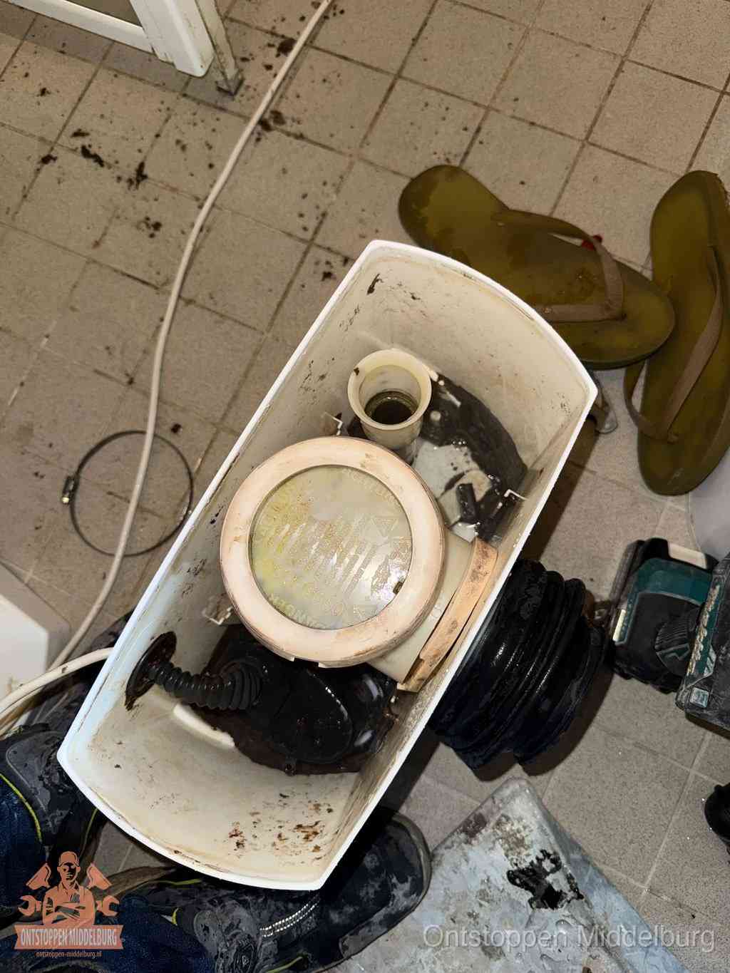 Een gedemonteerde toiletpot op de vloer met ontstoppingsveer en gereedschap ernaast, het verwijderen van een vastzittend voorwerp uit de wc in Middelburg